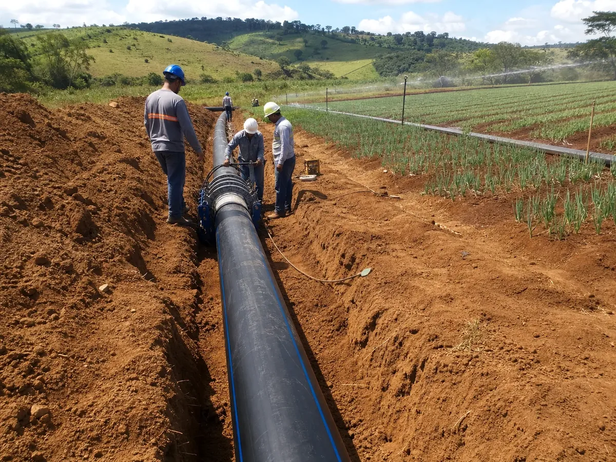 Obra rural de irrigação com tubo PEAD em vala, trabalhadores e área agrícola ao fundo